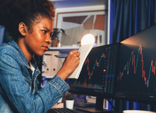 Concentrated young African woman trader writing analysis, looking on screen with stock market chart at high profit. Concept of exchange investment online in trading application on pc. Tastemaker.