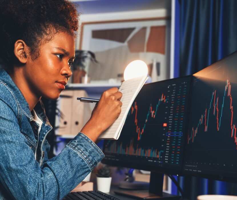 Concentrated young African writing analysis stock market screen. Tastemaker. Concentrated young African woman trader writing analysis, looking on screen with stock market chart at high profit. Concept of exchange investment online in trading application on pc. Tastemaker.